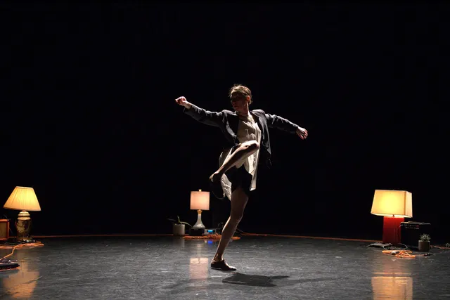 A female dancer wearing a white dress and blazer dancing with arms outstretched in a ring of orange extension cords and warmly lit lamps.
