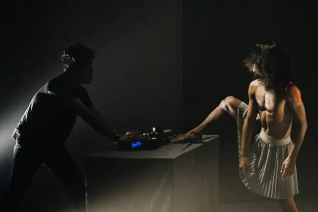 A shirtless person wearing a pleated skirt lifts their leg onto a keyboard behind controlled by a silhouetted figure on a black stage.