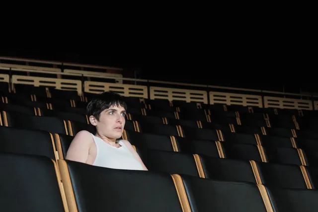 A woman wearing a white tank top sitting in a theater alone, staring at the stage with wide eyes.