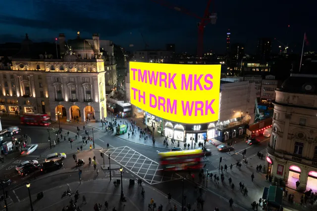 a large projection on a bright yellow screen in a town square reading TMWRK MKS, TH DRM WRK