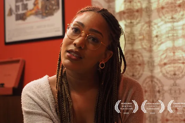 A Black woman wearing glasses and long braids looks toward the camera with head cocked in a room with red walls and patterned drapes.