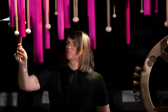 a white person in a black tshirt stands on stage with a mallet gesturing to play one hanging down from above