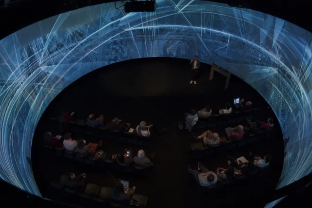 a group of people sit inside a 360 degree panoramic screen listening to a presentation. The screen has complex architectural imagery projected on it.