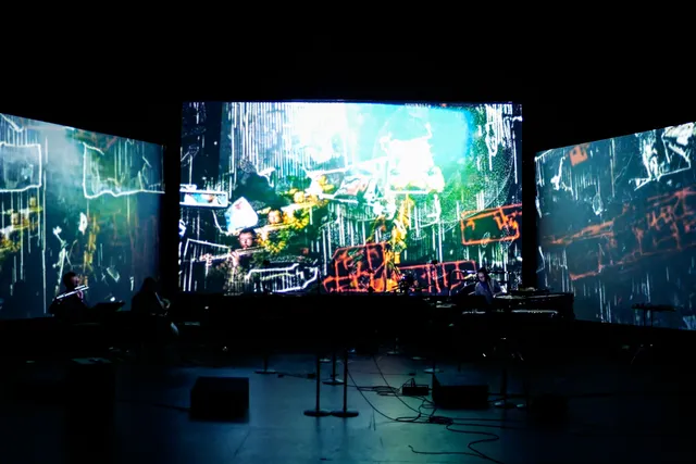 three screens with projections in the background of a band in the foreground of a black box studio.