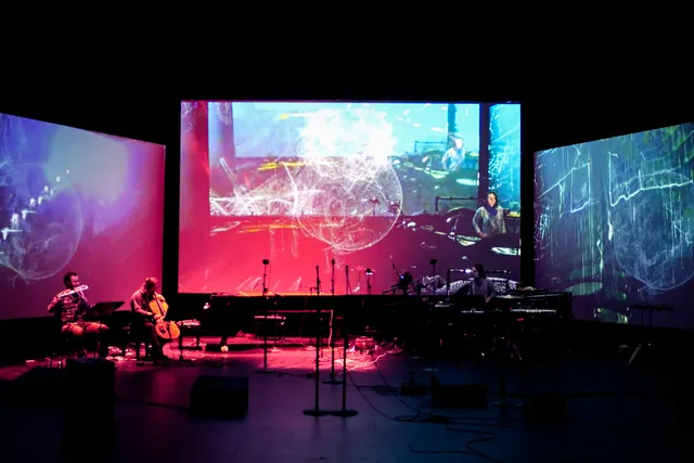 three large screens with projections in a black box studio with musicians performing.