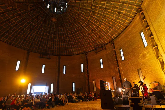 a crowd of concert-goers sits in chairs on hamster bedding in Troy's historic gasholder house while watching a five piece band.