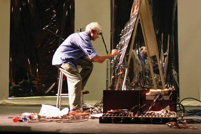 A man seated on a stole hunched over an easel surrounded by discarded wires and sound equipment. 