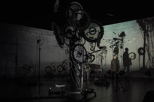 A man casting a long shadow standing near a sculpture of gears and wheels in a grey room. 