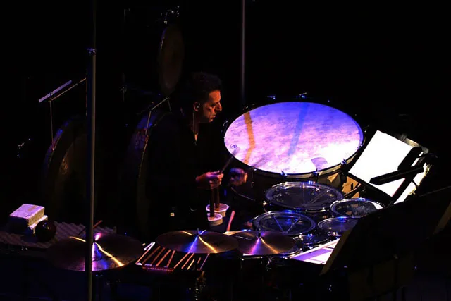 A man playing the drums washed in purple light on a dark stage. 