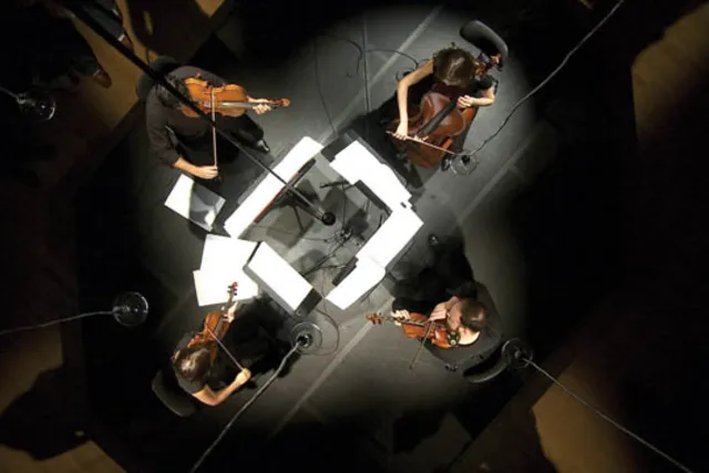An above shot of Mivos Quartet arranged in a square playing on a black stage. 