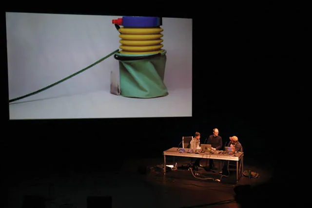 Three people seated behind a cluttered desk on a dark stage. A large projection screen behind them shows an image go an abstract green yellow and blue sculpture. 