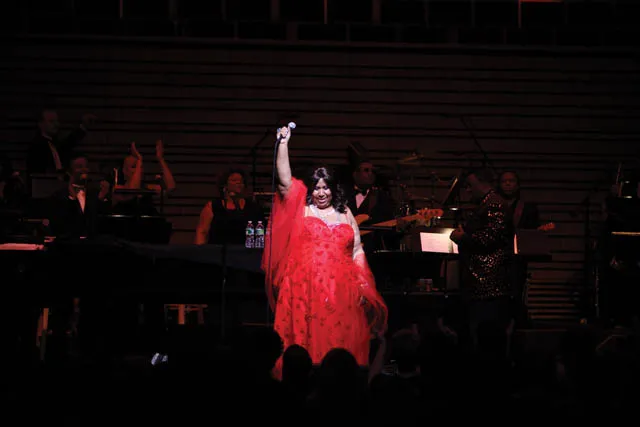 Aretha Franklin wearing a red gown and holding a microphone over her head on a dark stage. 