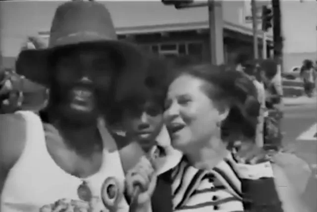A Black man wearing a sun har and white woman in the streets at a festival in 70s era clothing. 