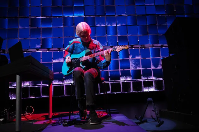 Ellen Arkbro seated playing electric guitar washed in blue and red light. 