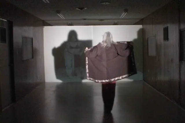 A woman creates a shadowy opening her jacket with back to viewer against a screen. 