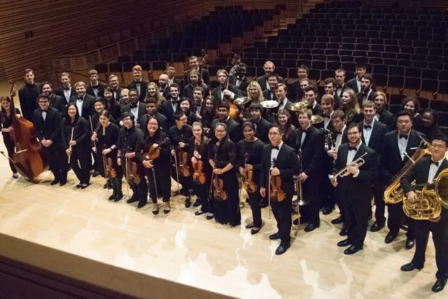 A student orchestra wearing black performance attire posing for a picture on stage of concert hall at EMPAC. 