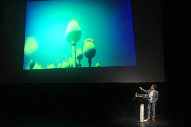 Mel Y. Chen in discussion gesturing at a large projection of blue and green plant buds.