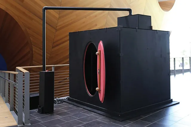 A back box with an oval red door and tubing extending out of the stop set up on the mezzanine floor set against the wooden hull of the concert hall. 