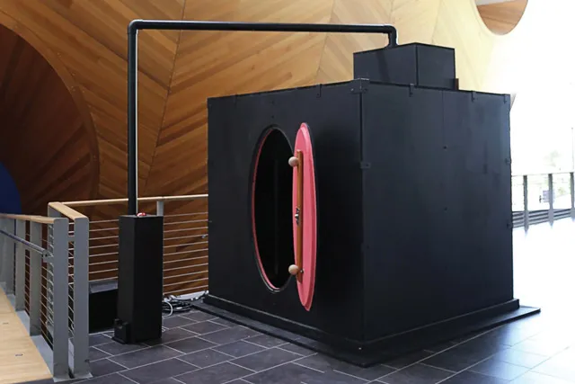 A back box with an oval red door and tubing extending out of the stop set up on the mezzanine floor set against the wooden hull of the concert hall.