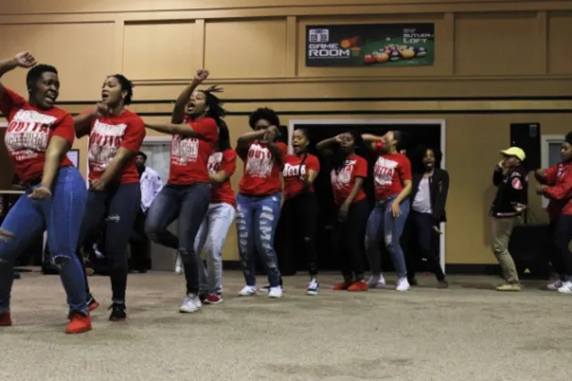 11 Black woman wearing red t-shirts and jeans dancing joyfully doing a set routine in a line around a room with beige walls and floors. 