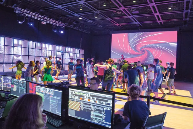 A crowd of people dancing on a yellow dance floor in front of a projection of a pink and purple swirl.
