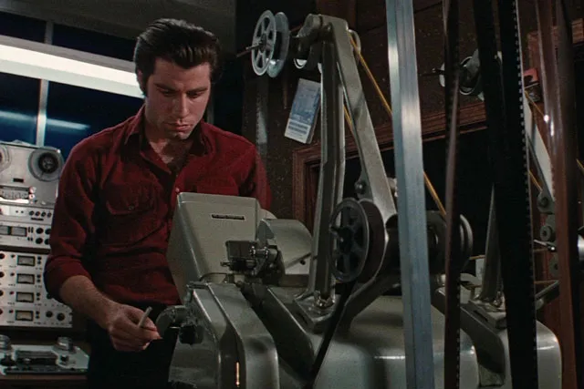 John Travolta standing behind a projector in a 1970's/80's projection booth.
