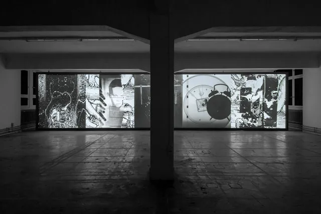 two screens with projections of colleges of various items, including an alarm clock and the face of a young boy in a subterranean room.