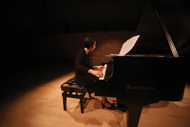 An asian woman with a bobbed haircut playing a grand piano on the dimly light concert hall stage. 