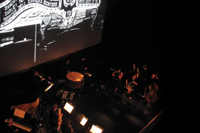 A pit orchestra in dim light playing at the foot of a large movie screen, showing a vintage black and white marquee. 