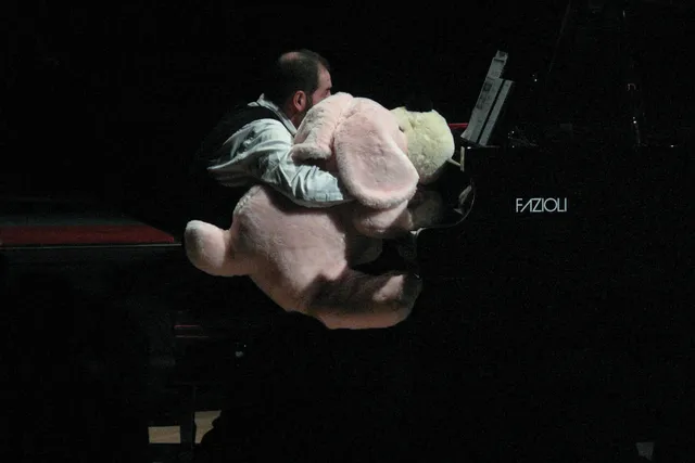 Luciano Chessa seated at a piano holding a large pink stuffed animal dog. 