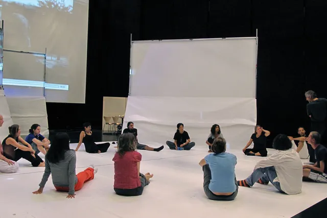 A  group of fourteen dancers dressed causally for rehearsal seating in a circle in a white floor listening intently to a speaker who is standing with arms gesturing outward.
