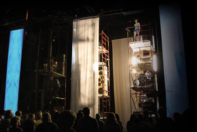 Musicians on scaffolding with each member of the band on different levels playing a concert to a crowd on a staged draped in sheer white fabric.