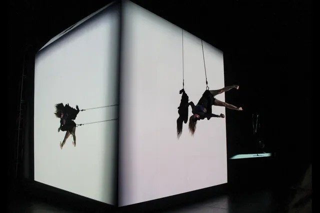 A woman suspended by a cable harness seemingly defying gravity while dancing on a white cube.