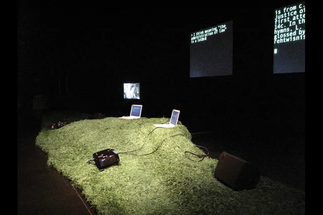 Two laptops sitting on an artificial grassy hill in front of two screens projecting blocky random text in a black box studio