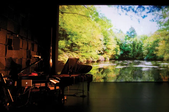 Two grand pianos among various wires, microphones, and recording devices in a black box studio in front of a projection of a tranquil green pond.