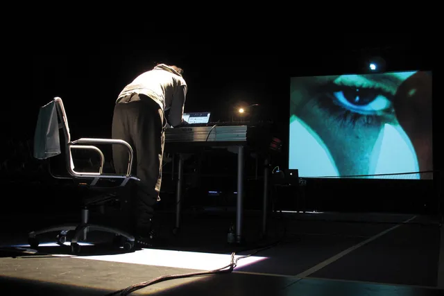 A person wearing gray sweatpants and sweatshirt standing hunched over a desk with backside to the viewer working on a computer controlling the projected image of a human eye.