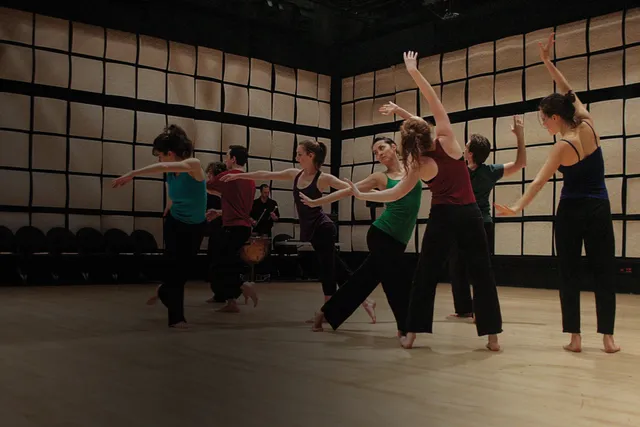 Eight dancers wearing jewel toned tops with black bottoms, moving with arms outstreched in a room with tan acoustic tiles on the walls.