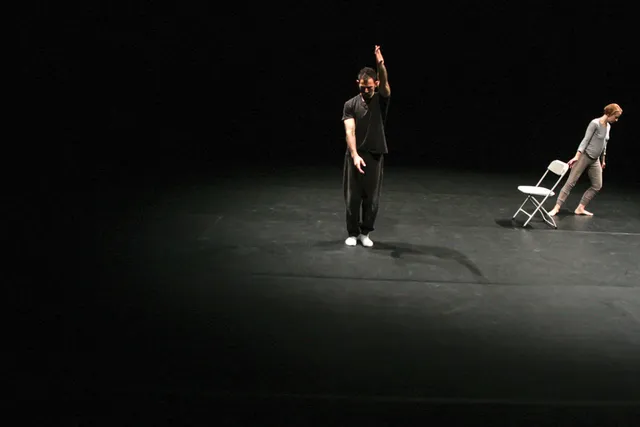 A male dancer wearing all black standing with one arm outstretched towards the ceiling as a female dancer wearing white drags a chair with body in a diagonal pose on a black stage.