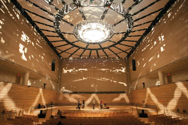 The EMPAC Concert Hall lit with spotty filtered light, mimicking the way light dapples through trees. A group of people stand on the stage among a few chairs.