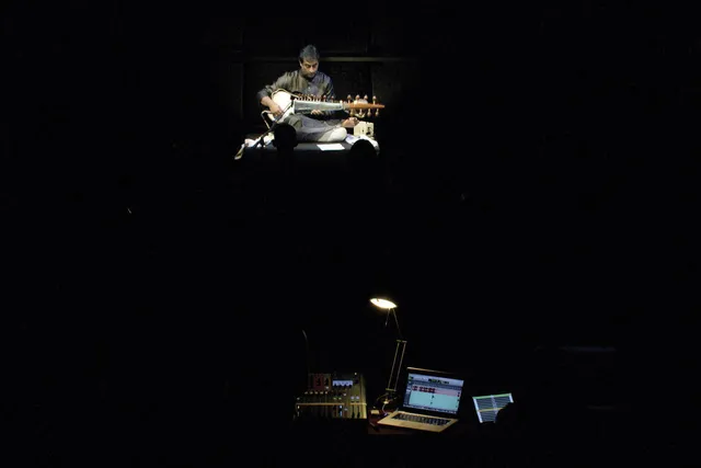 A man seated playing the sitar in a dark room, lit only by the sound panel in front of the stage.