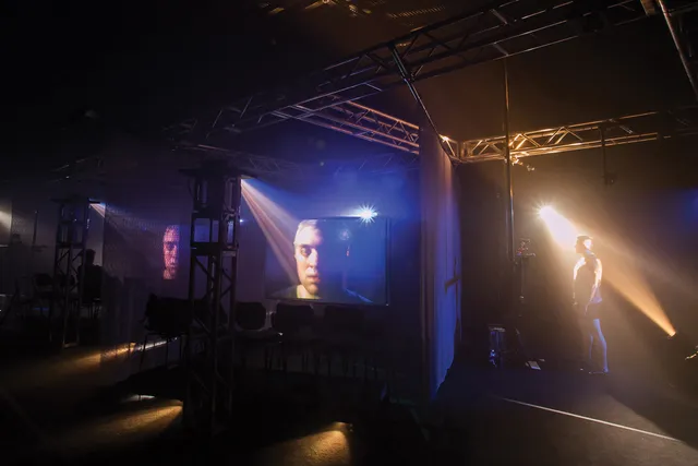 a male standing off to the right in a beam of theatrical light as his face is projecting on to two adjacent screens.