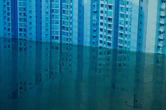 A blue high-rise apartment building with mean windows projecting on to a wall. A small puddle forming a reflection is on the floor where the wall meets the ground. 