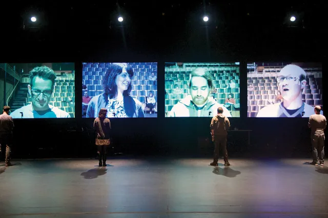 Four people standing on stage, back to the viewer looking at a projected image of themselves on a four screens in front of them.