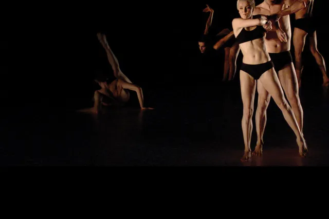 Five dancers wearing black underwear preforming on a black stage. A male dancer can be seen on the ground, back to the viewer with leg up as the rest are in a diagonal line.