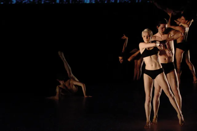 Six white dancers on dark stage dressed only in black underwear. Each person is in there own unique pose, including an individual on the floor with their leg extended and a woman standing in the front with arms twisted. 
