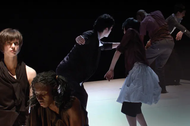 Six dancers wearing street clothes moving throughout the space diagonally. A white woman and a Black woman stand in the foreground wearing black draped dresses. The white woman Is looking off into the distance as the Black woman hunches, looking down.