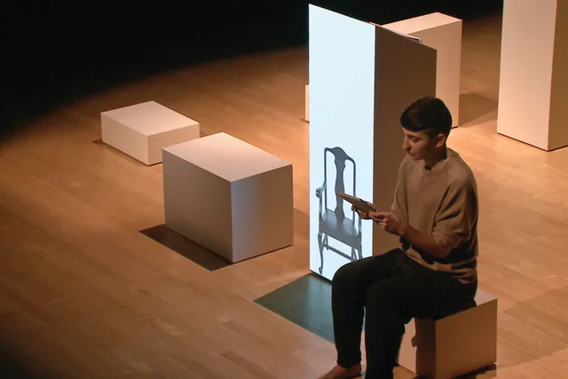 Gordon Hall sitting on white cube next to a projection of a chair on to a larger white cube 