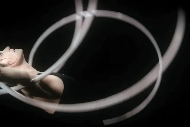 A white woman with dark hair arching her back and looking up with multiple white hoops spinning on her outstretched arms against a black background. 