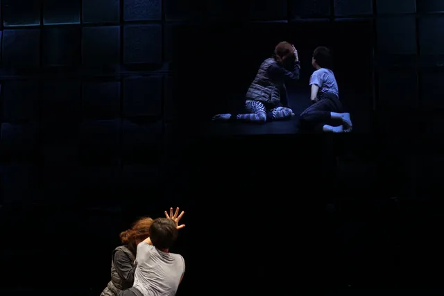 Two people in a black box studio reaching up towards a projection of themselves.