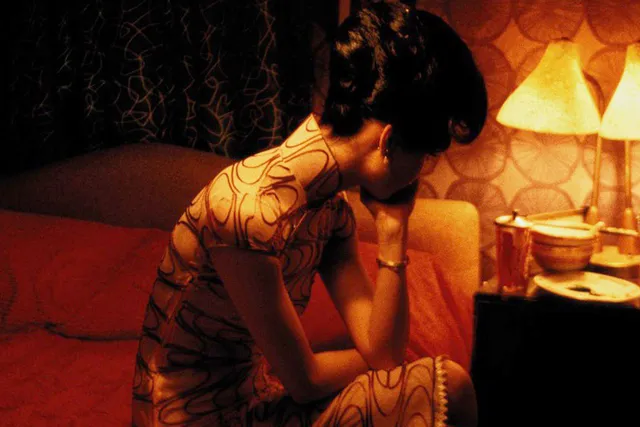 An asian woman wearing a Cheongsam sitting on a bed in an orange lit room with her head in her hand looking forlorn.